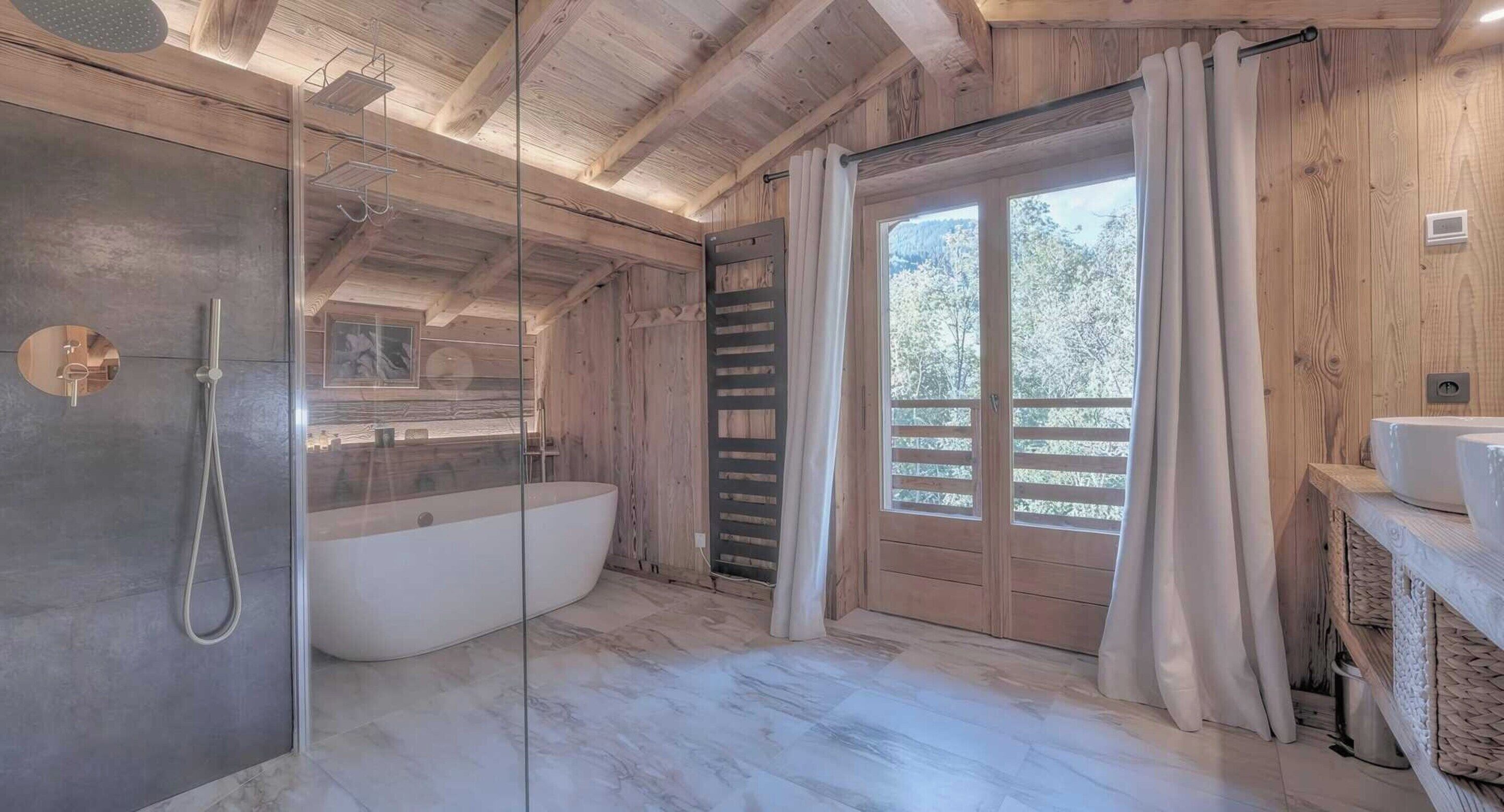A bathroom with a glass-enclosed shower, a freestanding bathtub, wooden walls, and a large window with a view of trees.