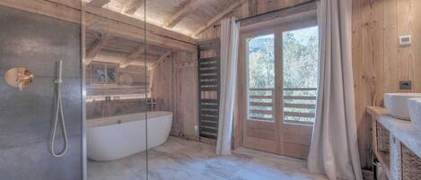 A bathroom with a glass-enclosed shower, a freestanding bathtub, wooden walls, and a large window with a view of trees.