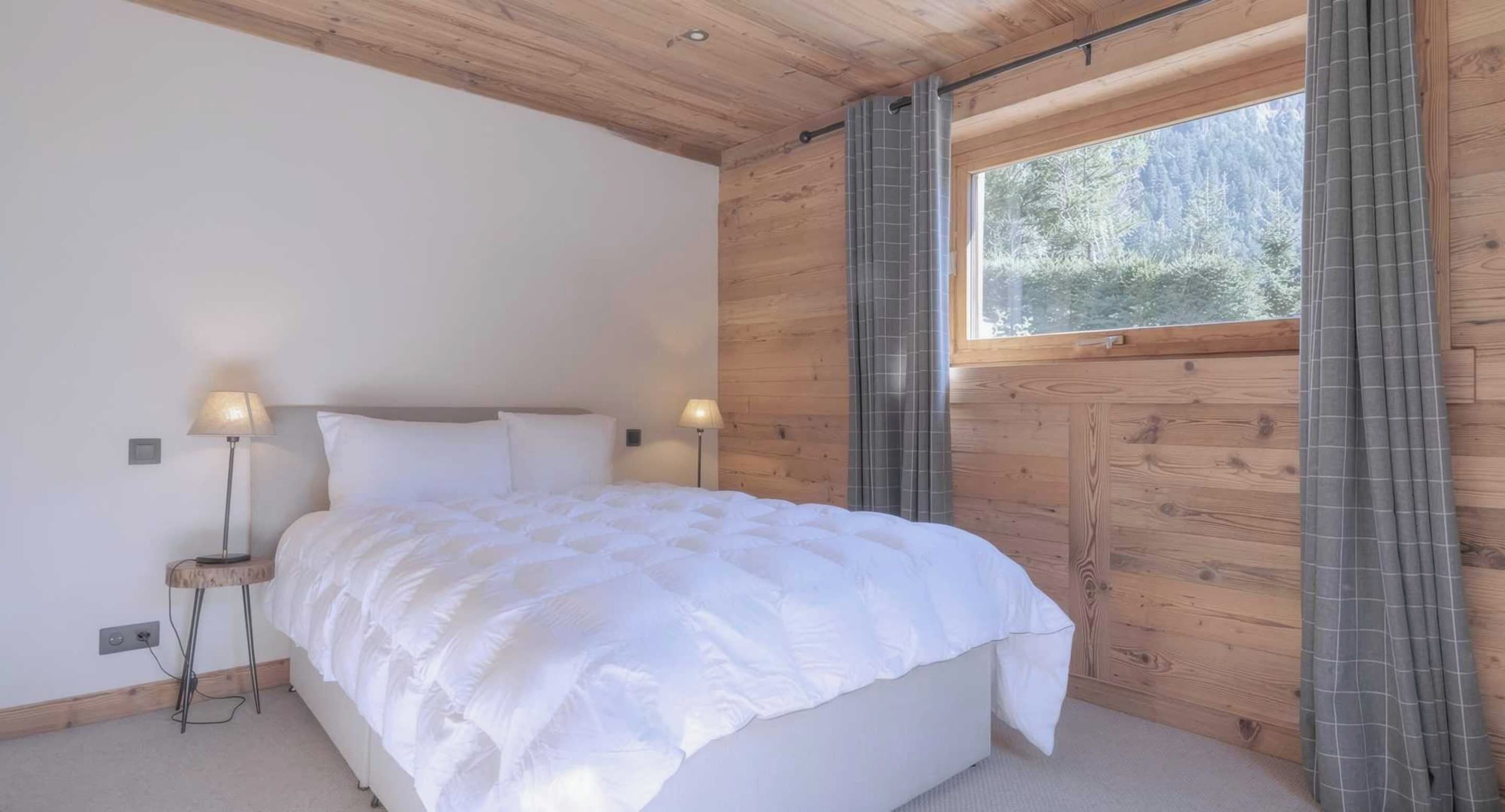 A bedroom with a bed, two bedside lamps, a window with curtains, and wooden walls.