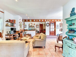 A spacious living room with a sofa, armchair, and a TV on a wooden stand. There are shelves with books and decorative items, a wooden door, and a wall with a clock and framed pictures. - Quietud by Interhome (Sta Cristina d'Aro)