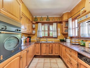 A kitchen with wooden cabinets, a stove, a sink, and a window with curtains. - Quietud by Interhome (Sta Cristina d'Aro)