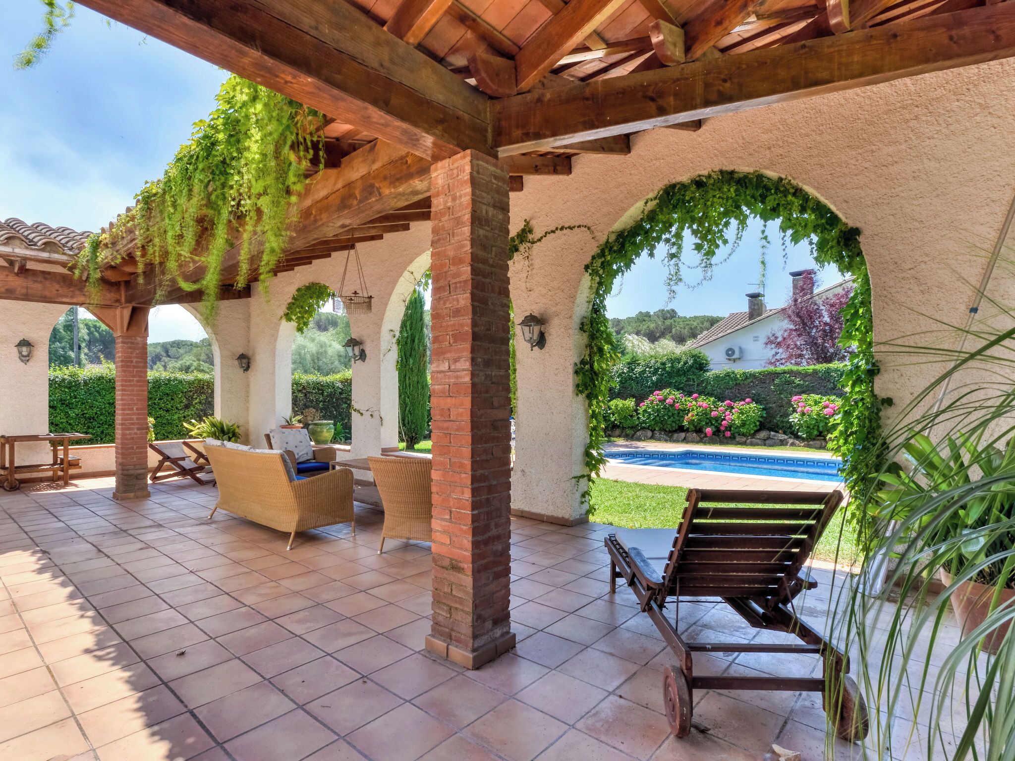 Ein überdachter Patio mit Gartenmöbeln, einem Pool und viel Grün.