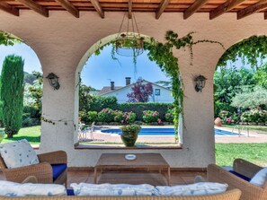 Un espace extérieur aménagé avec des meubles en osier, abrité par un auvent, offrant une vue sur une piscine et une maison en arrière-plan.