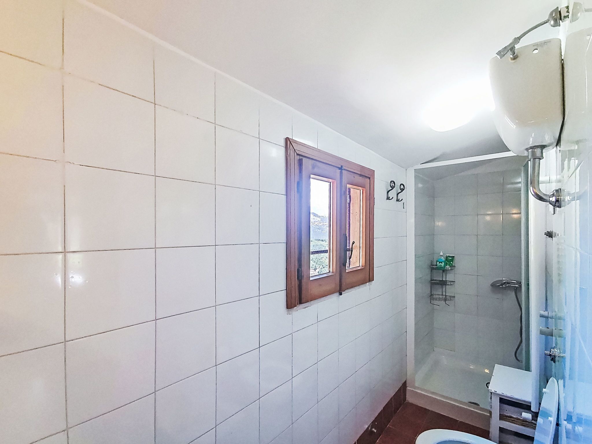 A bathroom with white tiled walls, a shower enclosure, a toilet, and a window with a wooden frame.