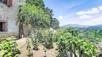 Un edificio in pietra con recinzione in legno, un giardino con diverse piante e vista su dolci colline lontane.