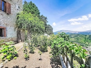 Un edificio in pietra con recinzione in legno, un giardino con diverse piante e vista su dolci colline lontane.