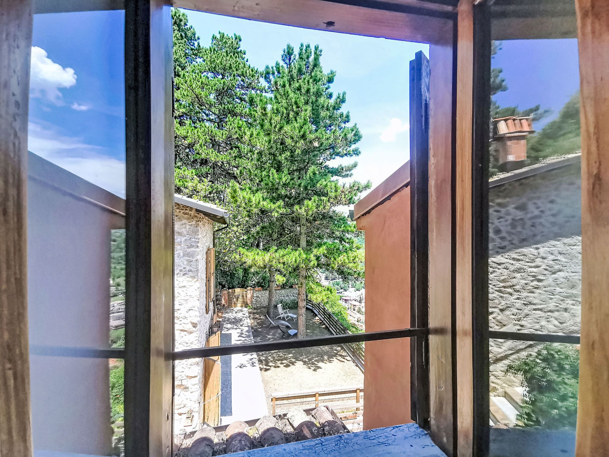 Blick durch ein Fenster auf ein Steinhaus mit einer Holztür, einer Steinmauer und einem Baum im Hintergrund.