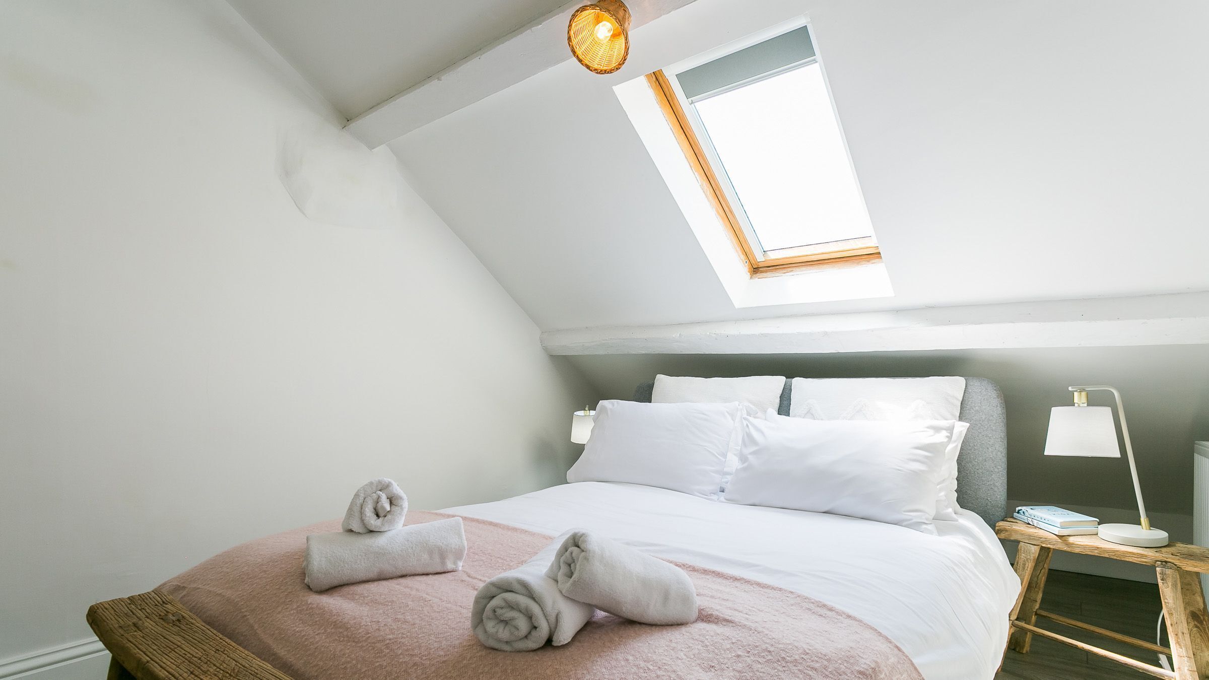 A bedroom with a skylight, a bed with white linens, a bedside table with a lamp, and a rolled towel on the bed.