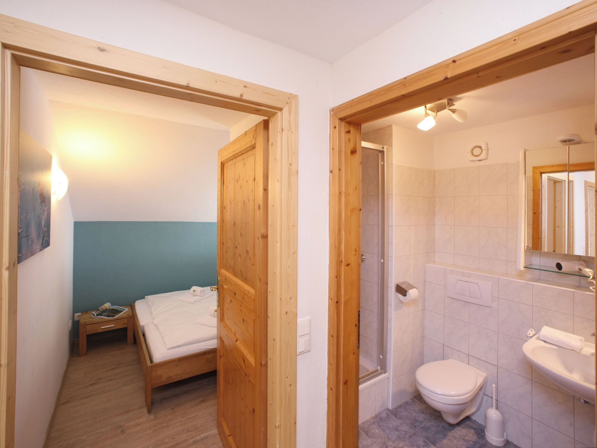 A bedroom with a wooden door, a bed, and a nightstand. Adjacent is a bathroom with a toilet, sink, and shower.