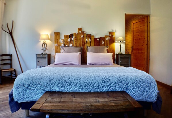 A bed with a blue textured cover, two bedside tables with lamps, a wooden headboard, and a wooden bench at the foot of the bed.