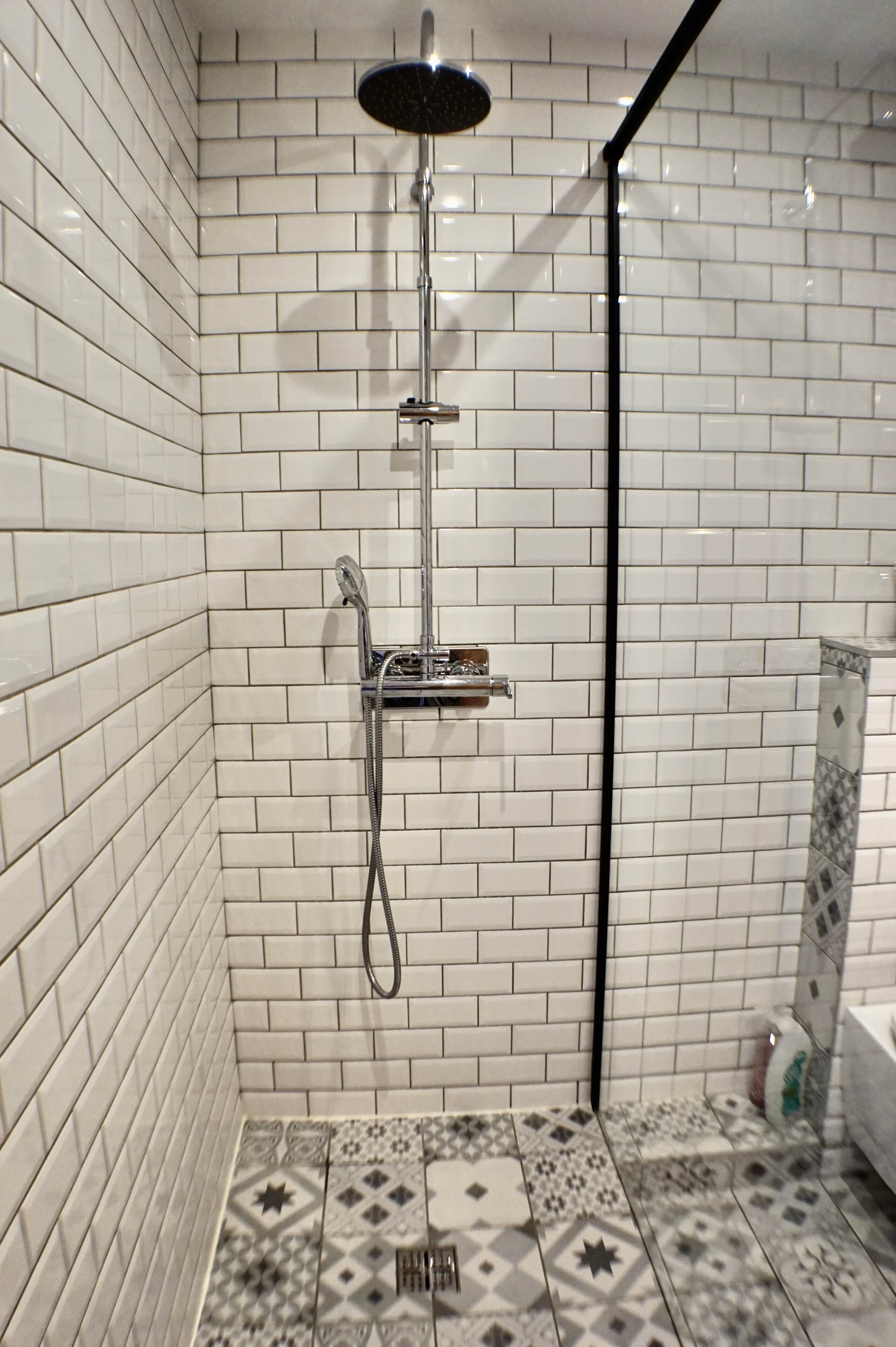 A shower with a handheld showerhead and a wall-mounted faucet, tiled with white and patterned tiles.
