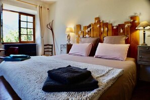 A bedroom with a large bed, a window with curtains, a wooden headboard, bedside lamps, and a chair.