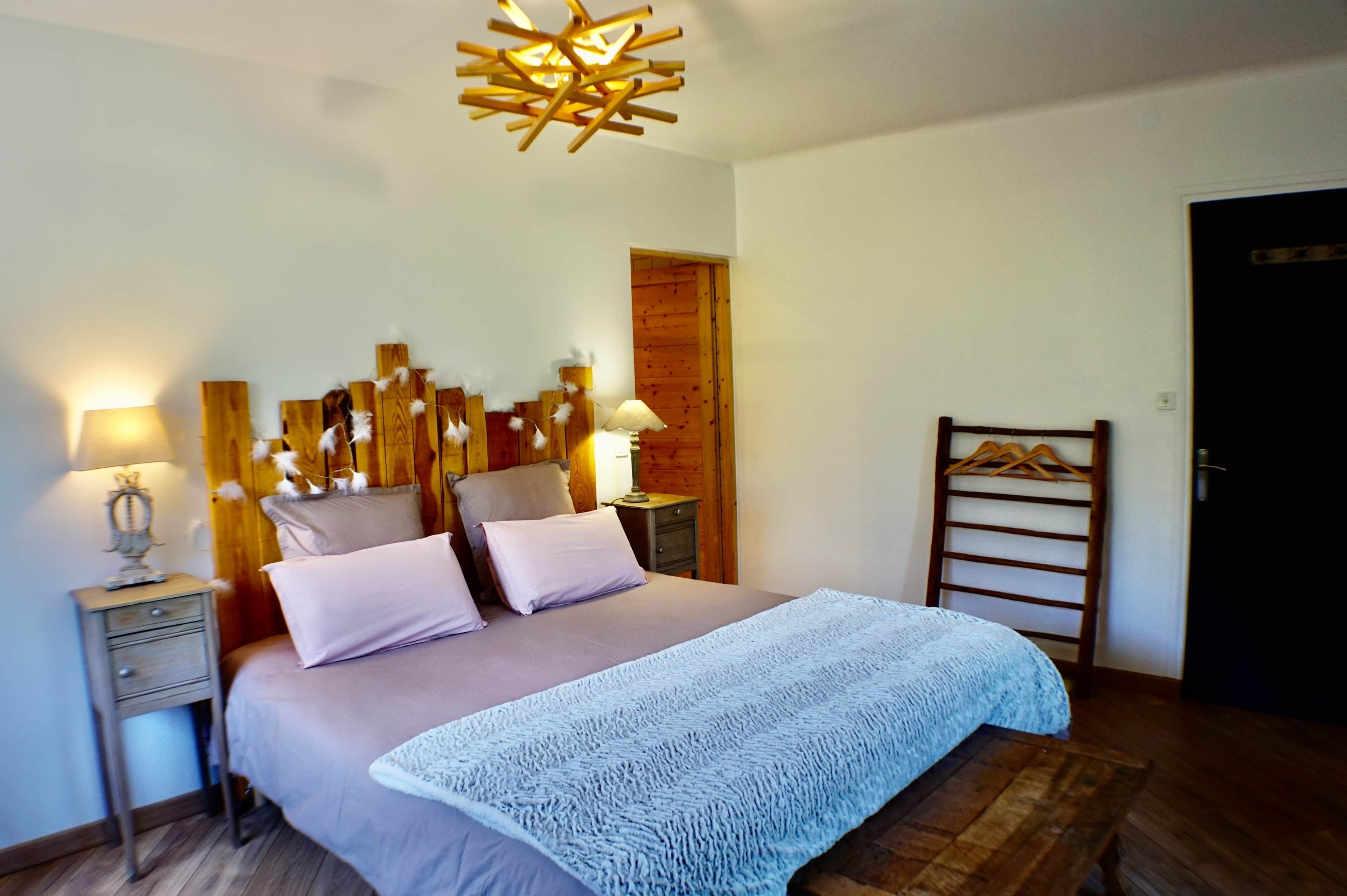 A bedroom with a bed, bedside tables, a hanging light fixture, a wooden headboard, a wooden door, and a wooden bench.