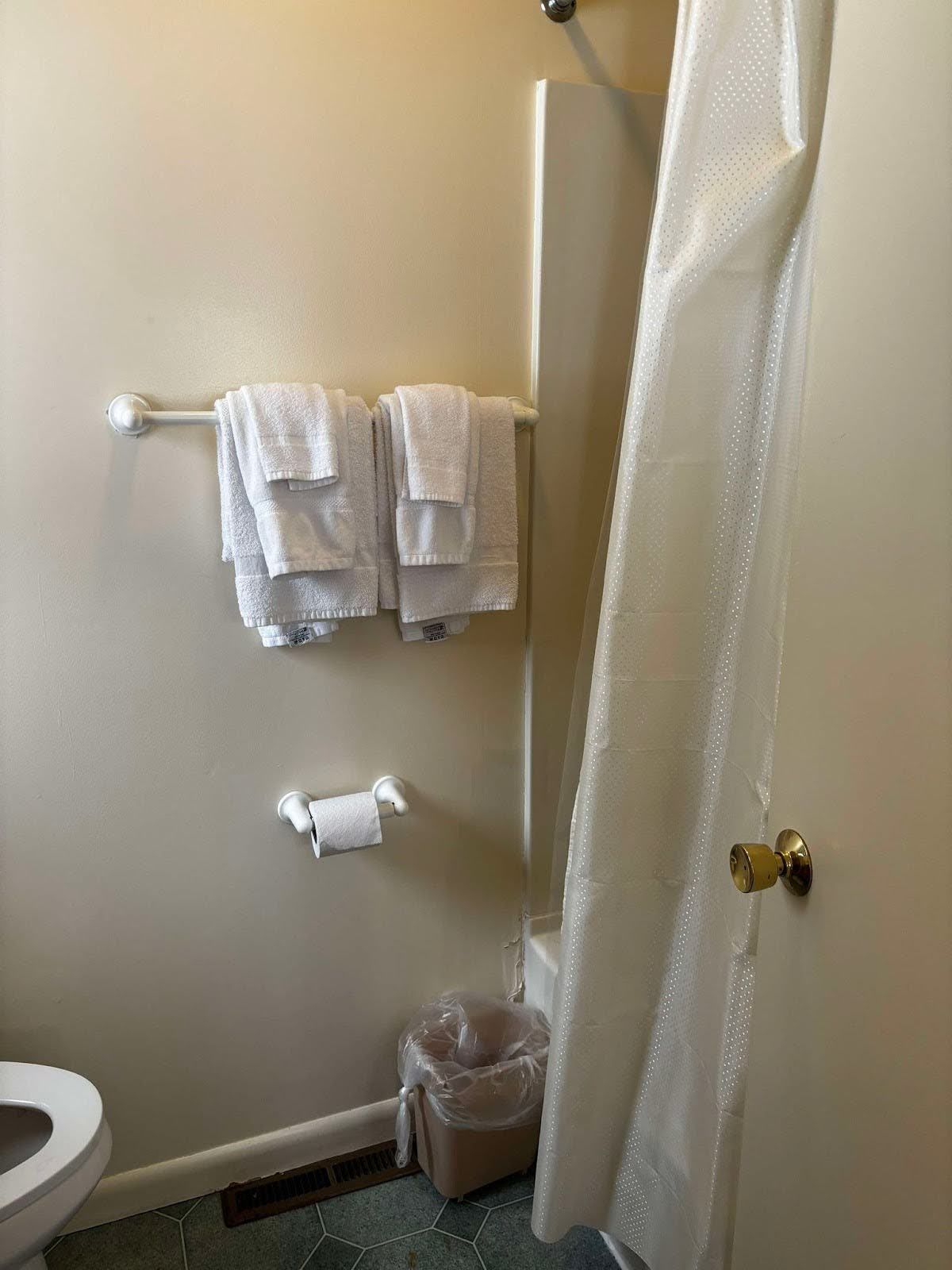 A bathroom with a toilet, a small trash bin, a towel rack with folded towels, and a shower curtain.