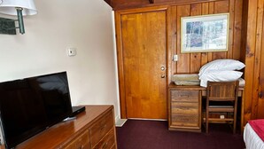 A wooden-paneled room with a television on a dresser, a bed with a red bedspread, a wooden chair, and a framed picture on the wall.