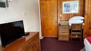 A wooden-paneled room with a television on a dresser, a bed with a red bedspread, a wooden chair, and a framed picture on the wall.
