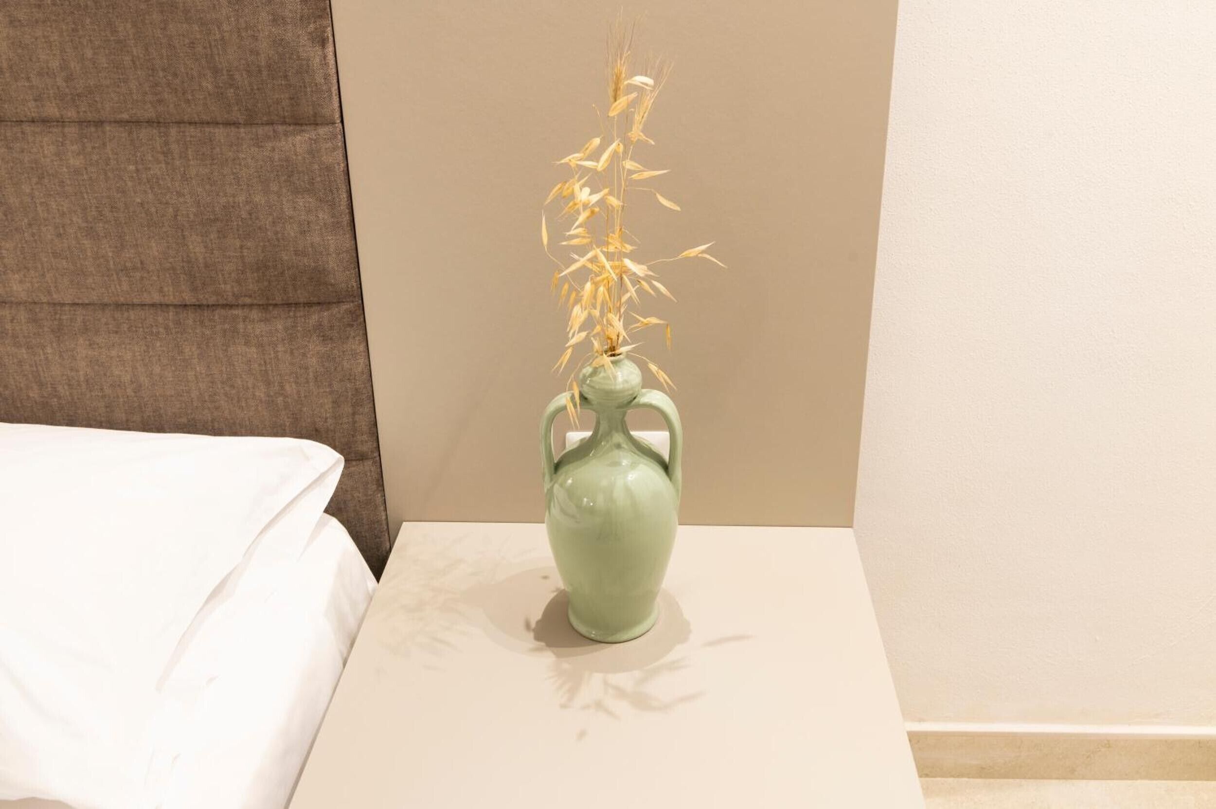 Un vaso verde con una pianta su una superficie bianca in una stanza con un letto e un muro marrone.