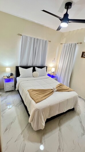 Une chambre d’hôtel avec un lit, deux tables de chevet avec des lampes, un ventilateur de plafond et des rideaux.