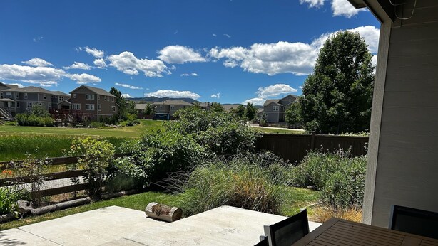 A patio with a concrete floor and wooden deck, overlooking a grassy area with a fence and a row of houses in the background. - Modern luxury home - great for families (Golden)