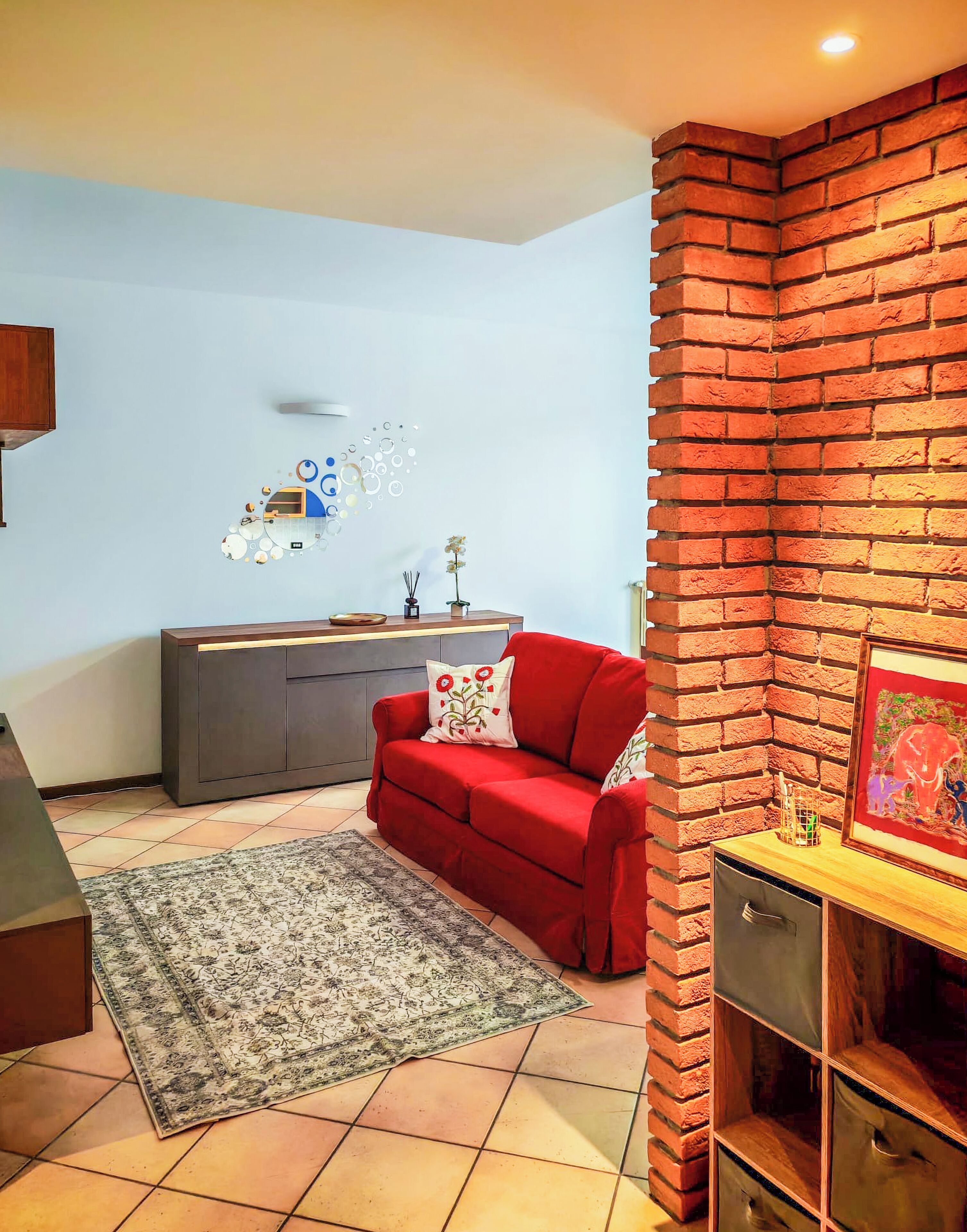Un salon avec un canapé rouge, un meuble gris, un tapis à motifs et un mur en briques.