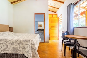 Un dormitorio con cama, techo de madera, baño visible a través de una puerta abierta y comedor con mesa y sillas.