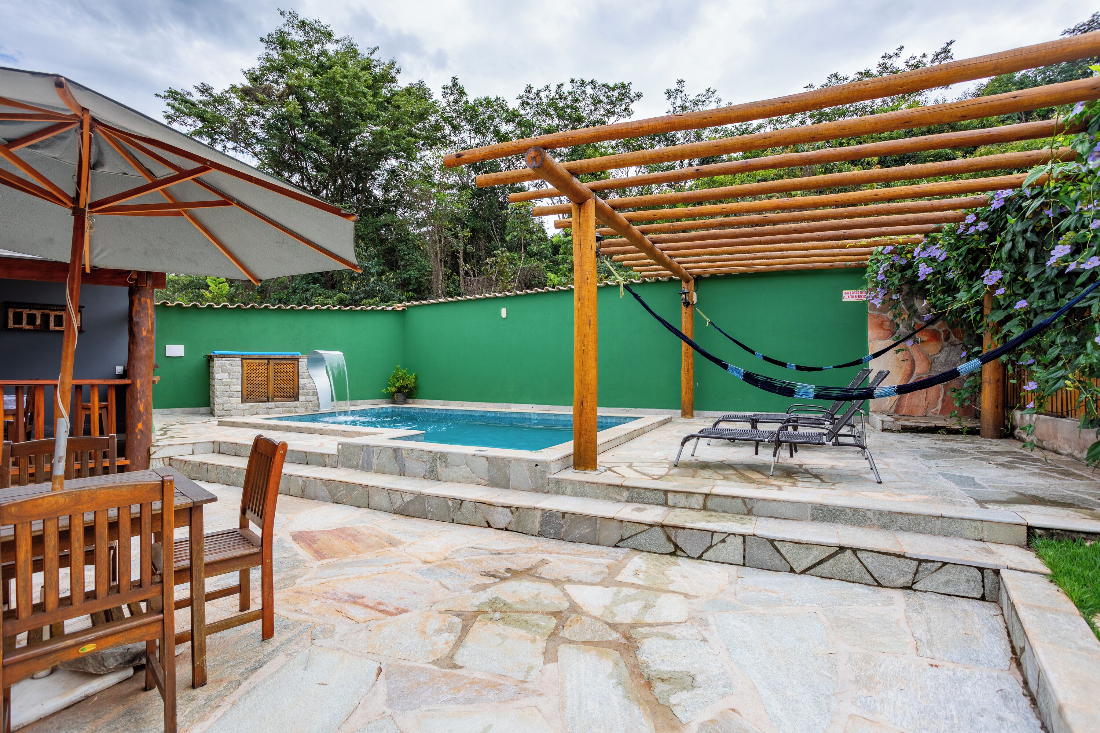 Uma área de piscina com um pergolado de madeira, uma rede e um espaço de estar com mesa e cadeiras.