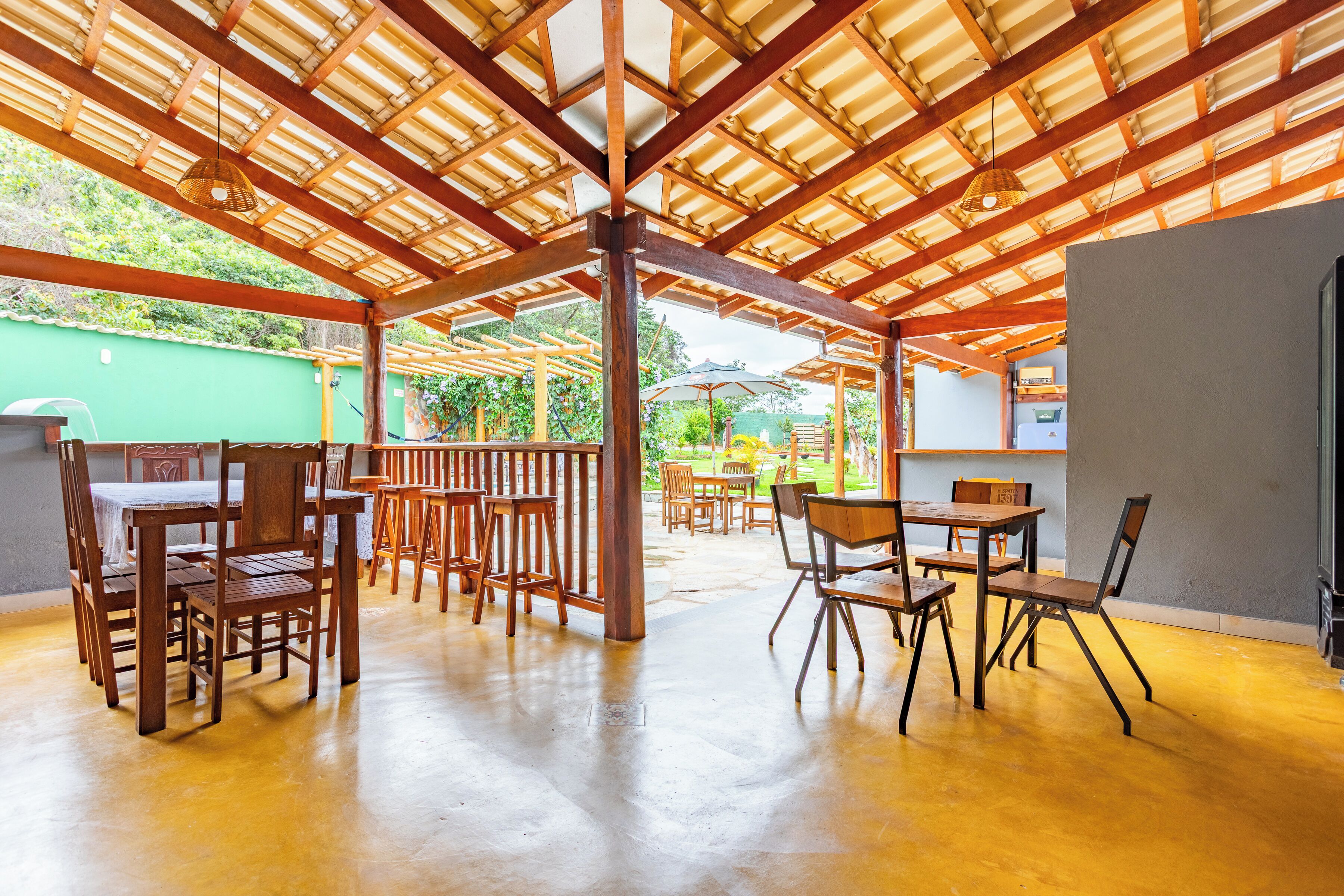 An outdoor patio area with wooden tables and chairs, a pergola structure, and a green wall in the background.