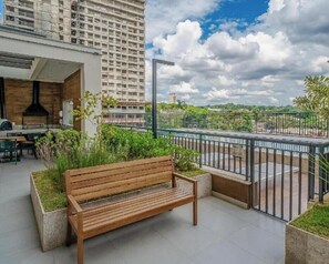 Eine Holzbank in einer begrünten Außenanlage mit Blick auf eine Stadtlandschaft und ein Gebäude im Hintergrund.