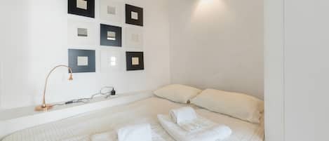 Un lit bien fait, recouvert d’une couvre-lit blanche à motifs géométriques, avec deux oreillers blancs et deux serviettes blanches soigneusement pliées.