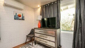 Una habitació amb paret de maó blanc, un televisor de pantalla plana sobre un moble de fusta, una cadira petita, un vas vermell i una finestra amb cortines.