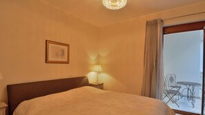 Une chambre d’hôtel comprenant un lit, une table de chevet, une lampe, un tableau encadré au mur et un balcon aménagé avec du mobilier d’extérieur.
