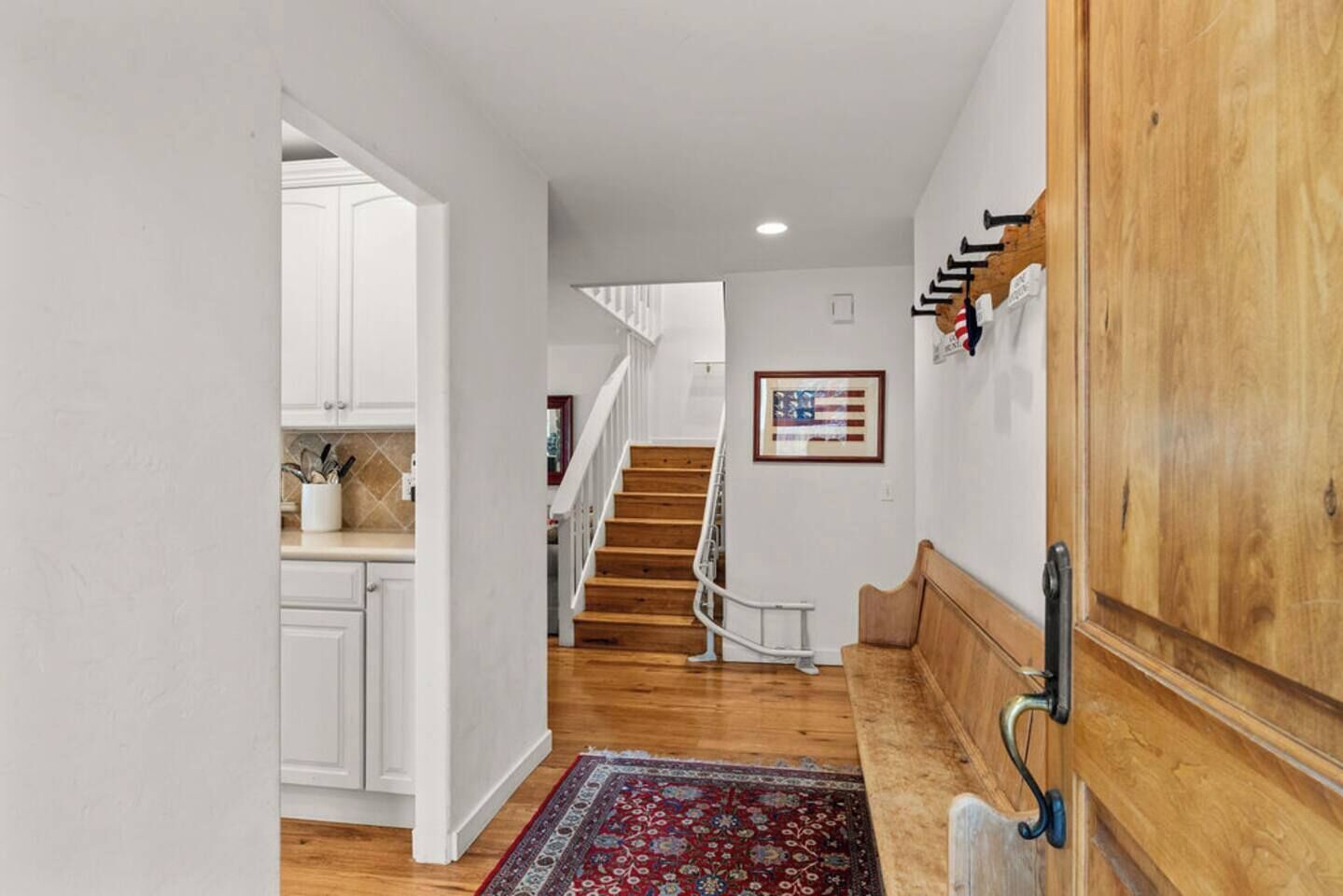 A hallway with wooden flooring, a red patterned rug, a staircase, and a kitchen area with white cabinets.