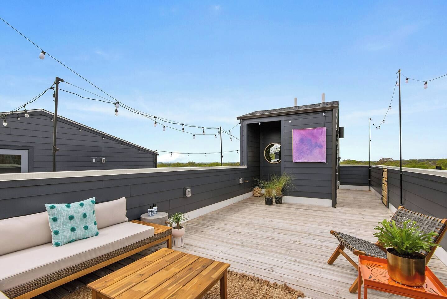 A rooftop deck with a sofa, chairs, a small table, string lights, and potted plants.