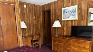 Una camera d'albergo con parete rivestita in legno, un letto con copriletto decorato, una scrivania con lampada, un comò con televisore e un quadro appeso al muro.