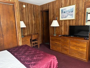 A hotel room with a wooden paneled wall, a bed with a patterned bedspread, a desk with a lamp, a dresser with a television, and a framed picture on the wall. - Cozy 1-bedroom hotel room, Hamilton - 311 Gilbreth (Hamilton)