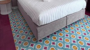 Uma cama com roupa de cama branca e duas almofadas vermelhas, cabeceira cinza e chão com padrão.