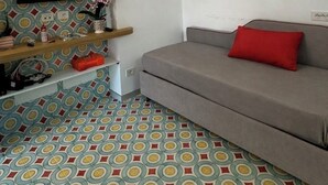 Quarto com sofá cinza, televisão de tela plana fixada em uma parede estampada, prateleira de madeira com diversos objetos e piso padronizado.