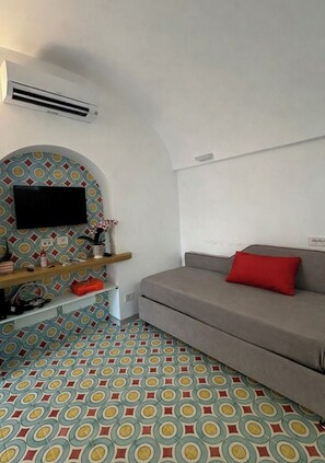 A room with a grey sofa, a flat-screen TV mounted on a patterned wall, a wooden shelf with various items, and a patterned floor. - Relais Totò:vacation home with authentic charm, inspired by  Naples (Campania)