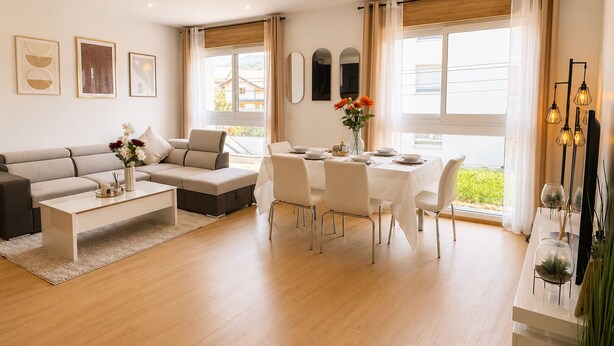 Una sala de estar moderna con un sofá, una mesa de centro, una mesa de comedor y sillas. Hay cuadros enmarcados en la pared, una ventana con cortinas y vistas a espacios verdes al exterior.