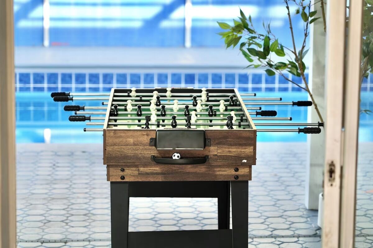 A foosball table with a wooden base and a digital scoreboard, placed in a room with a tiled floor and a pool in the background.