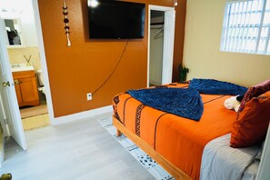 Una camera da letto con una coperta arancione, una televisione montata a parete e una finestra con persiane.