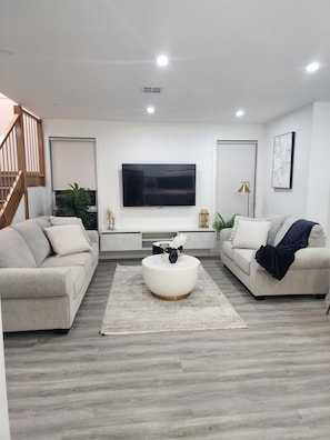 A modern living room with a flat-screen TV, two sofas, a coffee table, and a staircase.