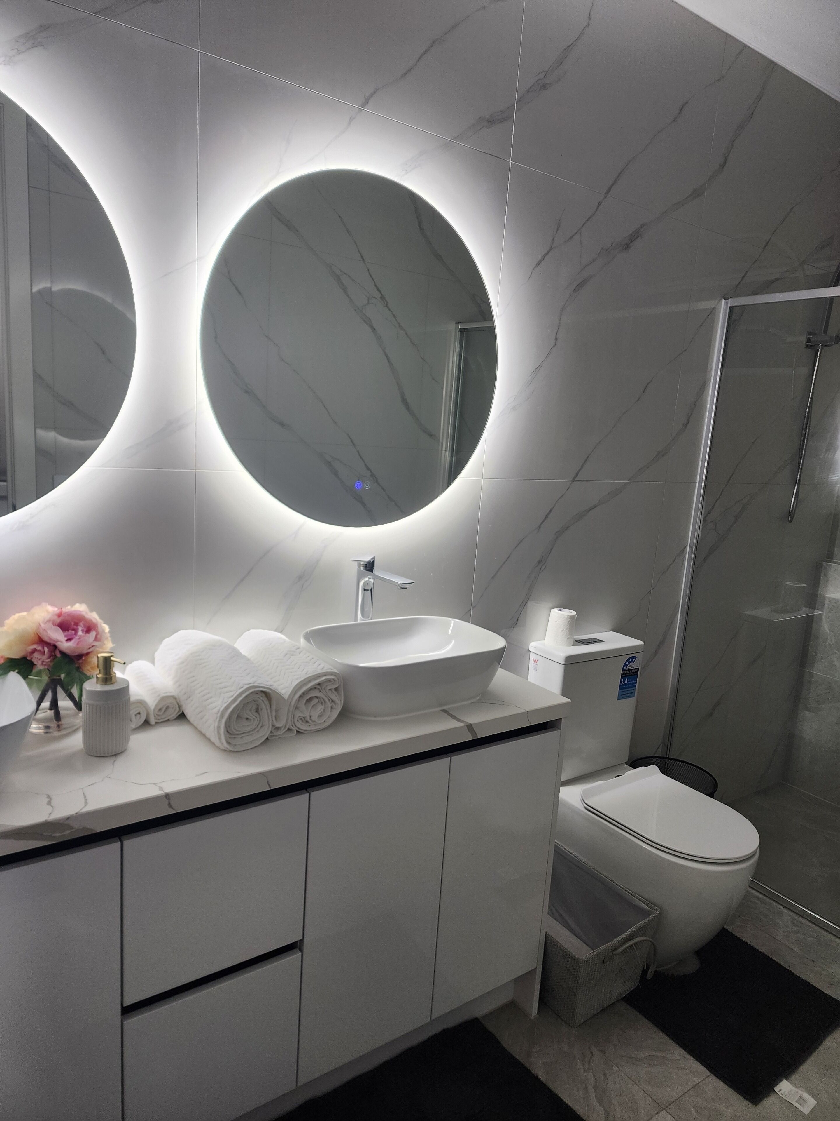 Ein modernes Badezimmer mit einem großen, ovalen Spiegel, einem weißen Waschbecken, einer Toilette und einer Dusche.