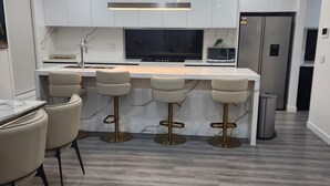 A modern kitchen with a marble countertop, four bar stools, a built-in oven, and a stainless steel refrigerator.