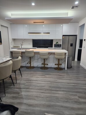 A modern kitchen with a marble countertop, four bar stools, a built-in oven, and a stainless steel refrigerator.