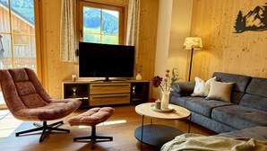 A wooden interior with a sofa, a chair, a TV, a lamp, and a view of a forest through a window. - Hideaway Chalets Loser - 1 (Altaussee)