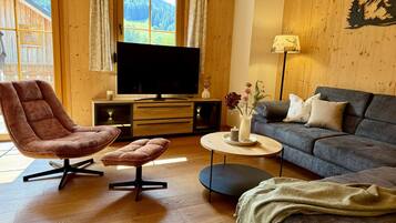 Un intérieur en bois comprenant un canapé, un fauteuil, un téléviseur, une lampe et une vue sur une forêt par la fenêtre.