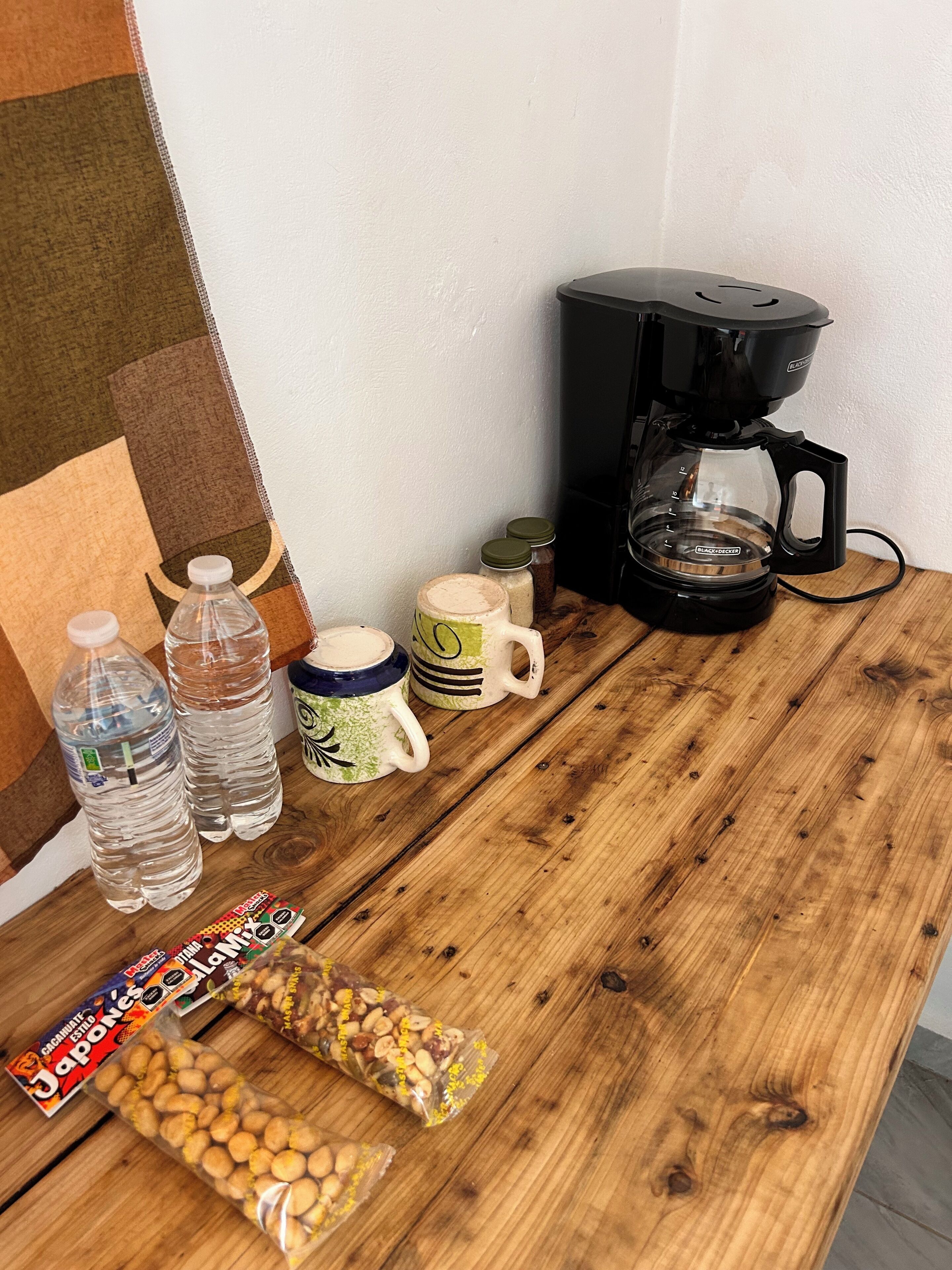 A wooden table with a coffee maker, water bottles, mugs, and snack packages.