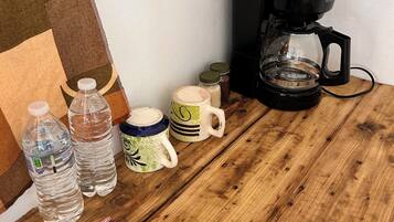 A wooden table with a coffee maker, water bottles, mugs, and snack packages.
