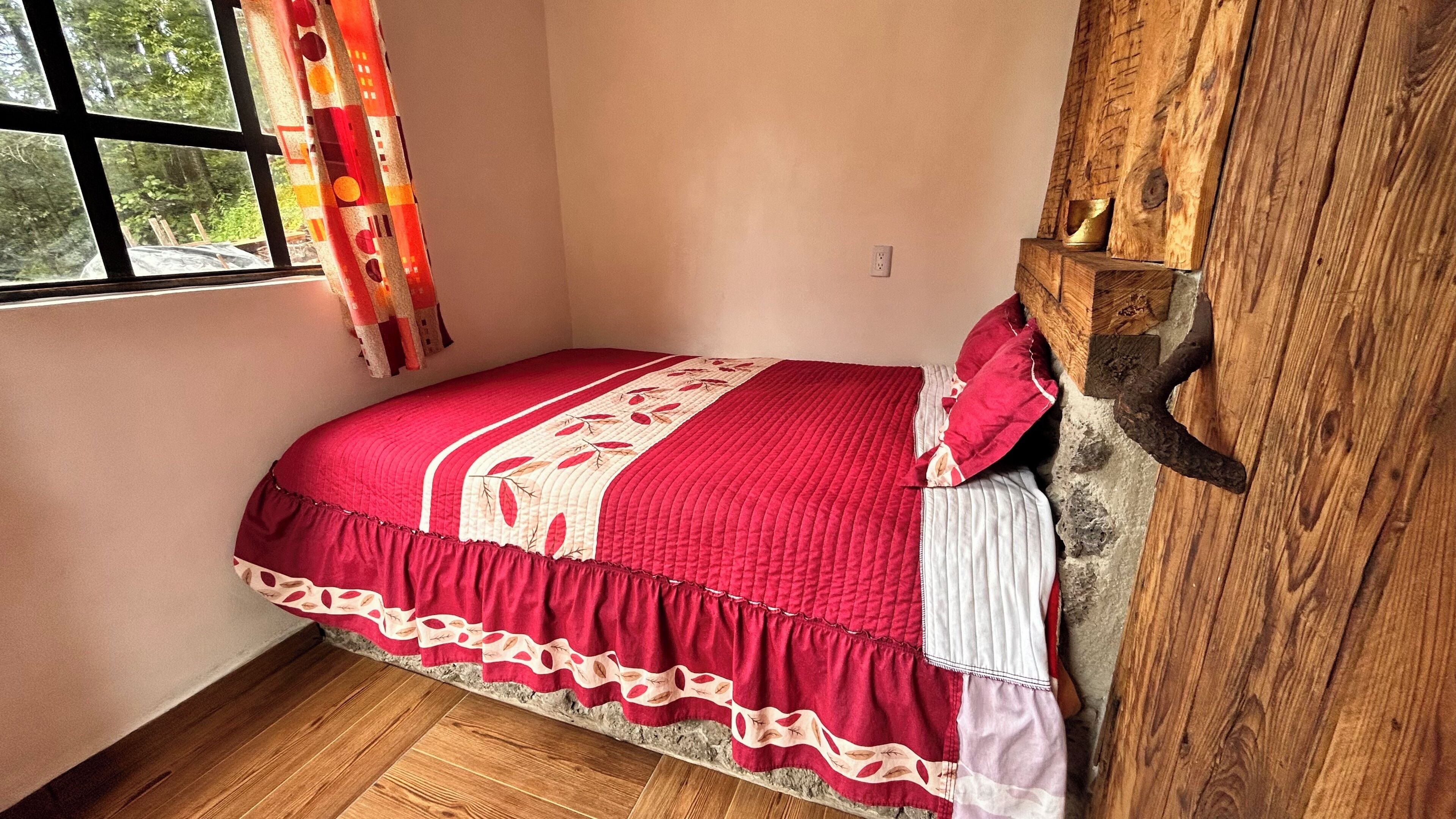 Une chambre avec un plancher en bois, une couvre-lit rouge et une fenêtre avec des rideaux.